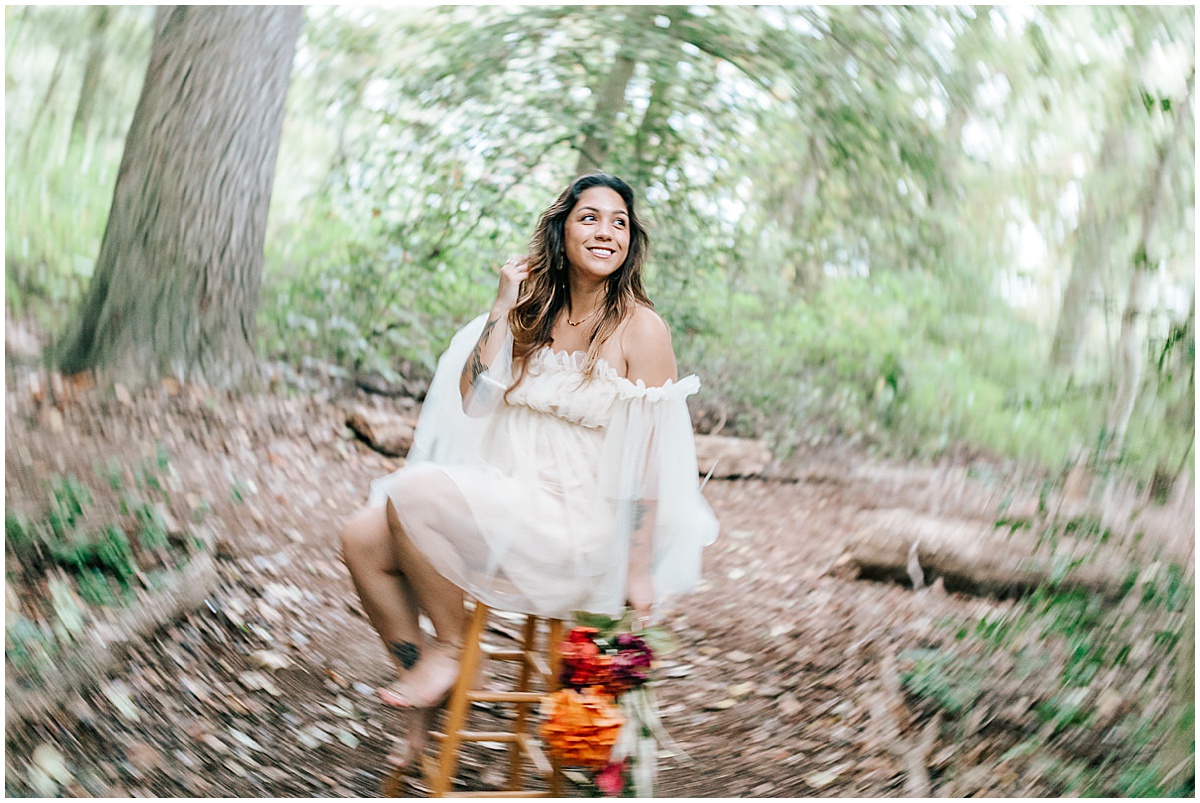 outdoor dreamy ethereal portraits in the woods in smyrna Delaware with Kass taken with a prims lens for a blurry effect on the surroundings. Kass is holding a colorful cottagecore bouquet while sitting on a wooden stool